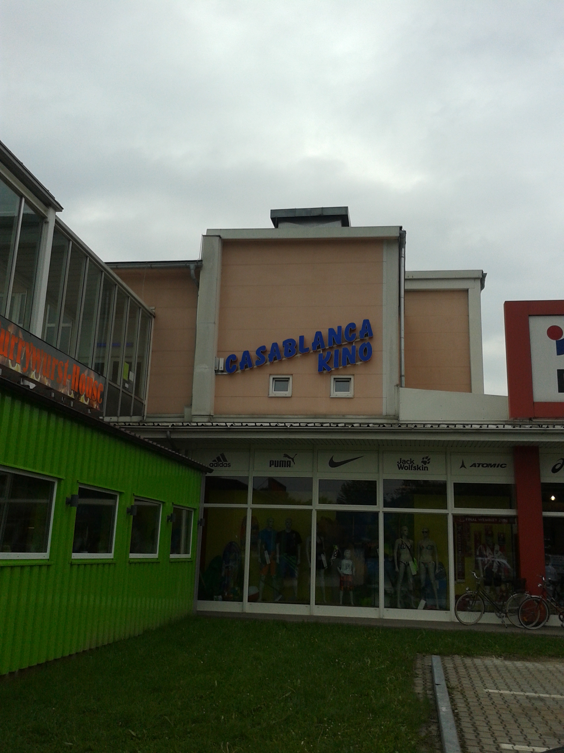 Zunächst sieht man eine Seitenansicht des Kinos. In der Mitte des Bildes liegt eine kleine Wiese, rechts kommt der Parkplatz ins Bild. Im Hintergrund ein Sportartikel-Geschäft, über dem das Kino eingerichtet ist.